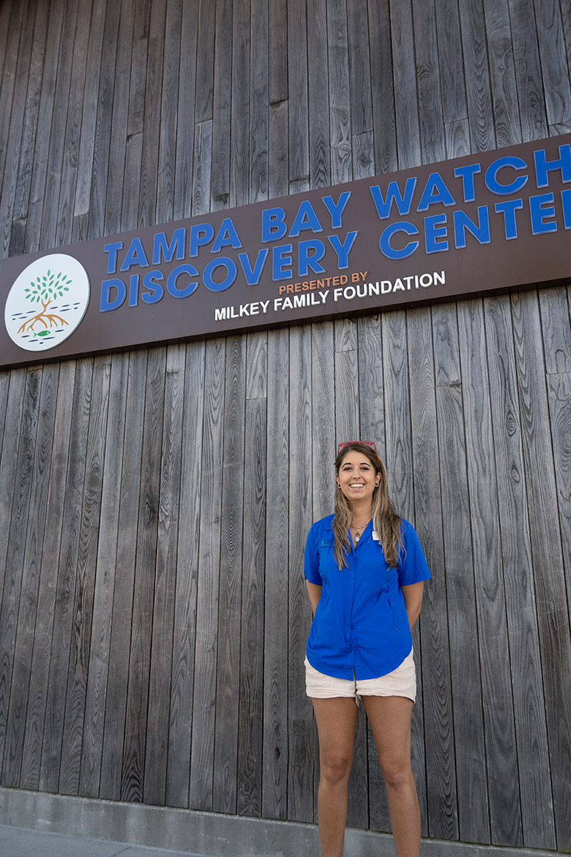 Woman stands in front of a wooden wall with signage that reads Tampa Bay Watch Discovery Center