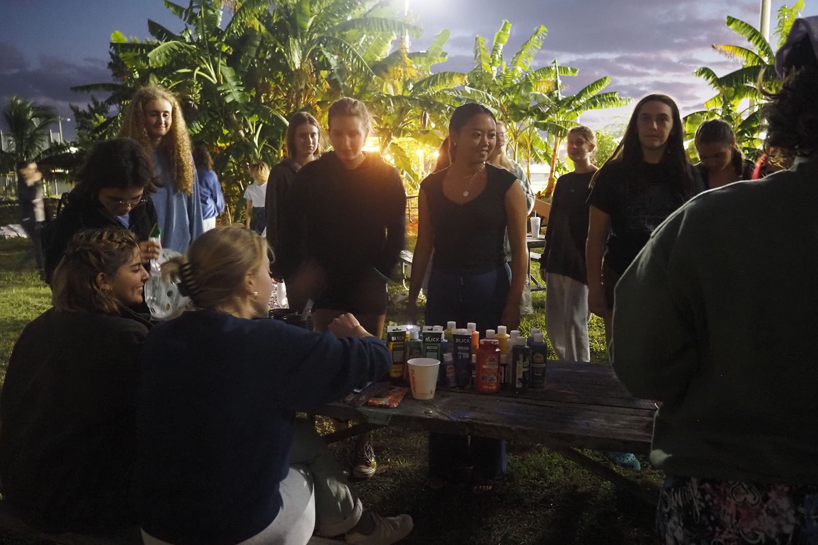 A group of students sitting and standing around a picnic table in the evening with banana trees in the background and paints on the table