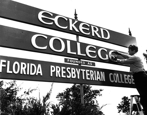 Black and white photo of man on a ladder adding the letter "e" to a sign that says "黄色短视频 founded as Florida Presbyterian College"